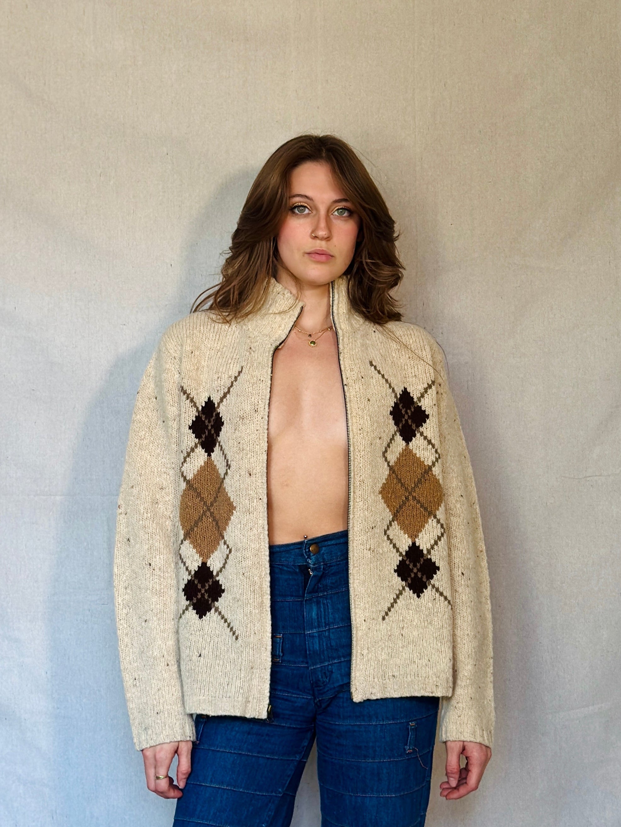 Person wearing a cream argyle sweater over a beige top with blue jeans against a plain background