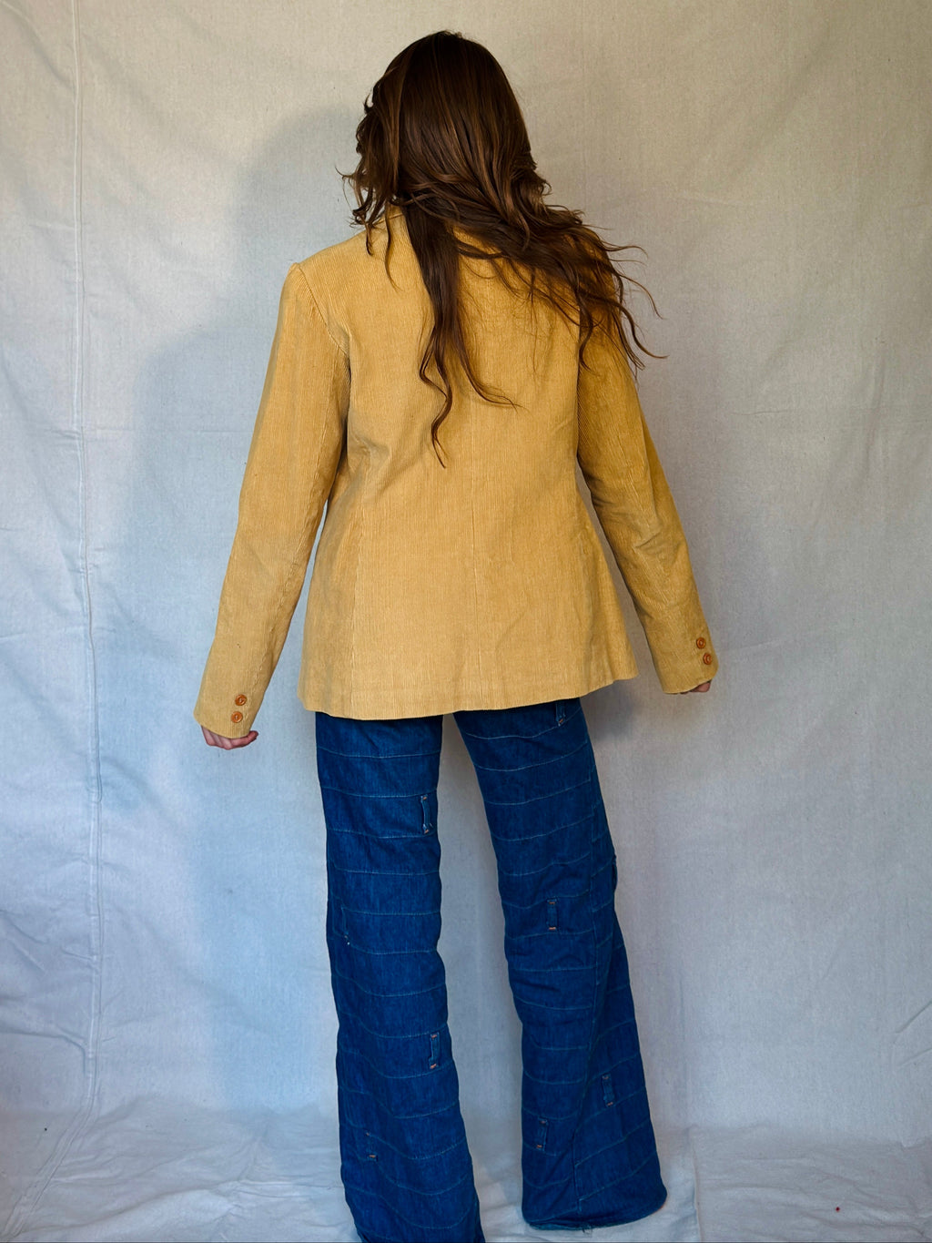 Person wearing a beige jacket and blue jeans against a plain background